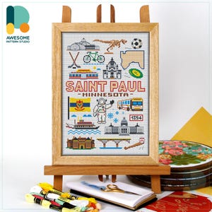 May include: A framed cross-stitch featuring a Saint Paul, Minnesota design. The artwork includes icons of local landmarks, a flag, and the text "SAINT PAUL - MINNESOTA". The frame is displayed on a wooden easel.