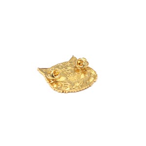 Puede incluir: Un pin dorado con forma de cara de gato, visto desde atr&aacute;s. El pin tiene dos cierres de alfiler y un dise&ntilde;o detallado. Los bordes son festoneados y el aspecto general es brillante y decorativo. El pin es probablemente de metal.