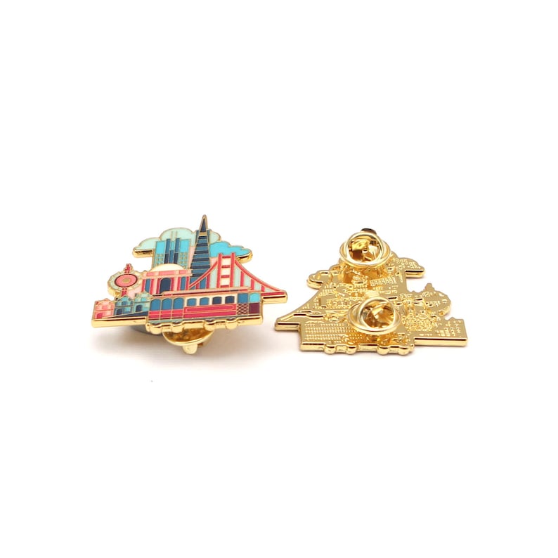 Op de afbeelding: Twee goudkleurige emaille pins. E&eacute;n pin toont een kleurrijke afbeelding van de skyline van San Francisco, inclusief de Golden Gate Bridge, een kabeltram en de Transamerica Pyramid. De andere pin toont de achterkant van de pin met de sluiting.