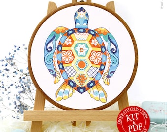 May include: A colorful cross-stitch pattern of a turtle with a geometric design. The pattern is displayed in a wooden embroidery hoop on a wooden easel. The image also includes a small tin with a cross-stitch pattern on it, a roll of blue fabric, and a set of embroidery floss.