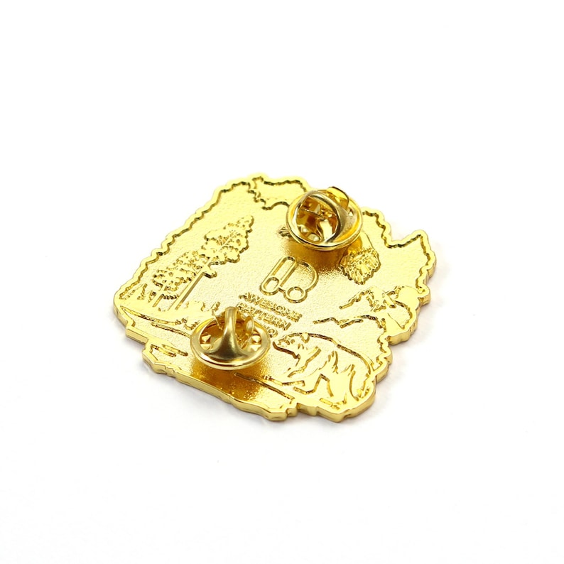 May include: A gold-colored pin shaped like a state, featuring a tree, binoculars, and a bear. The pin has two pin backs for secure attachment. The text "AMERICAN PATTERN" is visible on the pin.