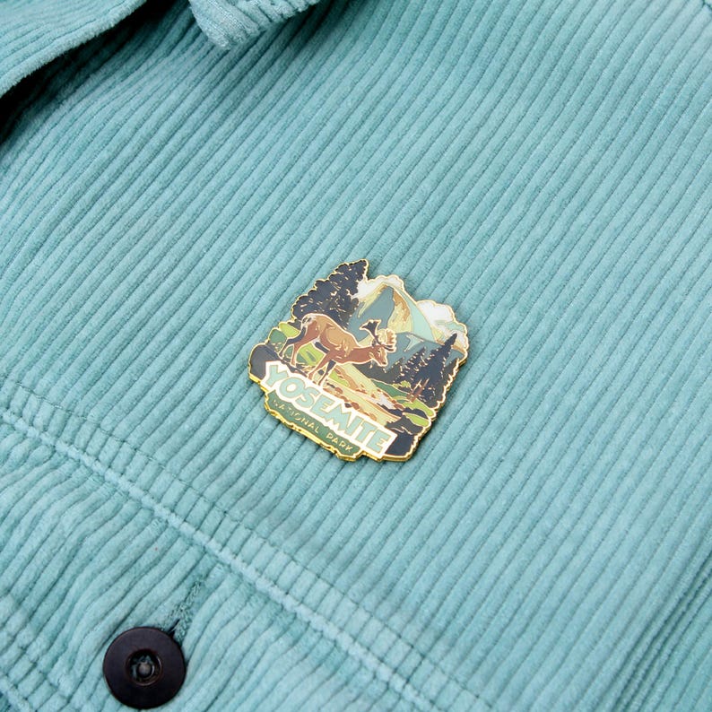 May include: A teal corduroy jacket with a Yosemite National Park pin. The pin features a deer, trees, and mountains with the words "YOSEMITE NATIONAL PARK" in a gold border.