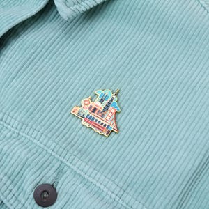 Op de afbeelding: Een goudkleurige emaille pin met een kleurrijke illustratie van een skyline, inclusief een brug en hoge gebouwen. De pin is bevestigd aan een lichtblauwe corduroy jas.