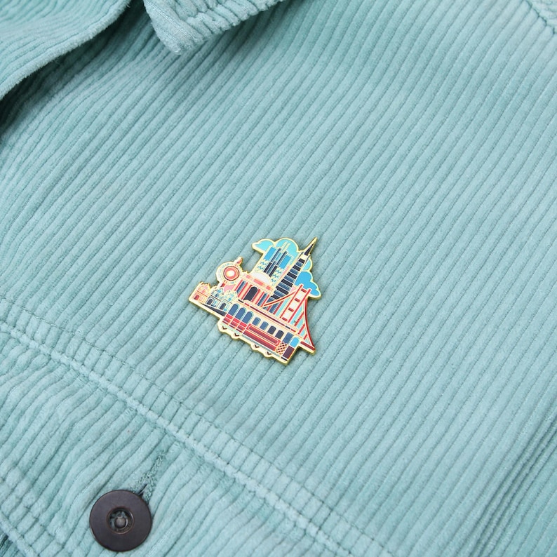 Op de afbeelding: Een goudkleurige emaille pin met een kleurrijke illustratie van een skyline, inclusief een brug en hoge gebouwen. De pin is bevestigd aan een lichtblauwe corduroy jas.