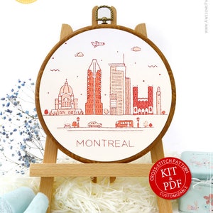 May include: A cross-stitch kit with a Montreal skyline design, presented in a wooden embroidery hoop on a small wooden easel. The design is in red and white, with the city's name below. The kit includes thread, scissors, and a pattern.