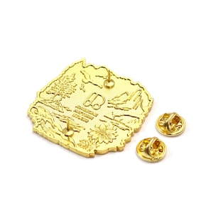 May include: A gold-colored metal pin in the shape of a state, featuring a detailed landscape design with trees, mountains, and a flower. The pin has two pin backs and the text "AWESOME PATTERN STUDIO".