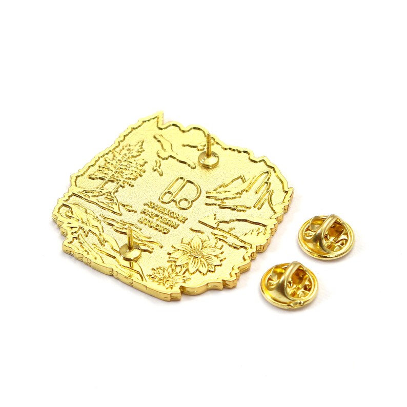 May include: A gold-colored metal pin in the shape of a state, featuring a detailed landscape design with trees, mountains, and a flower. The pin has two pin backs and the text "AWESOME PATTERN STUDIO".