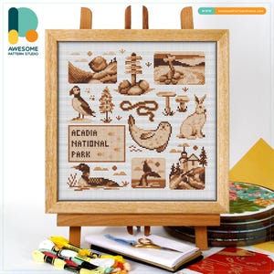 May include: A framed cross-stitch featuring Acadia National Park icons in shades of brown. The design includes a puffin, a rabbit, a seal, and other park elements. The text "ACADIA NATIONAL PARK" is also stitched into the design. The artwork is displayed on a wooden easel.