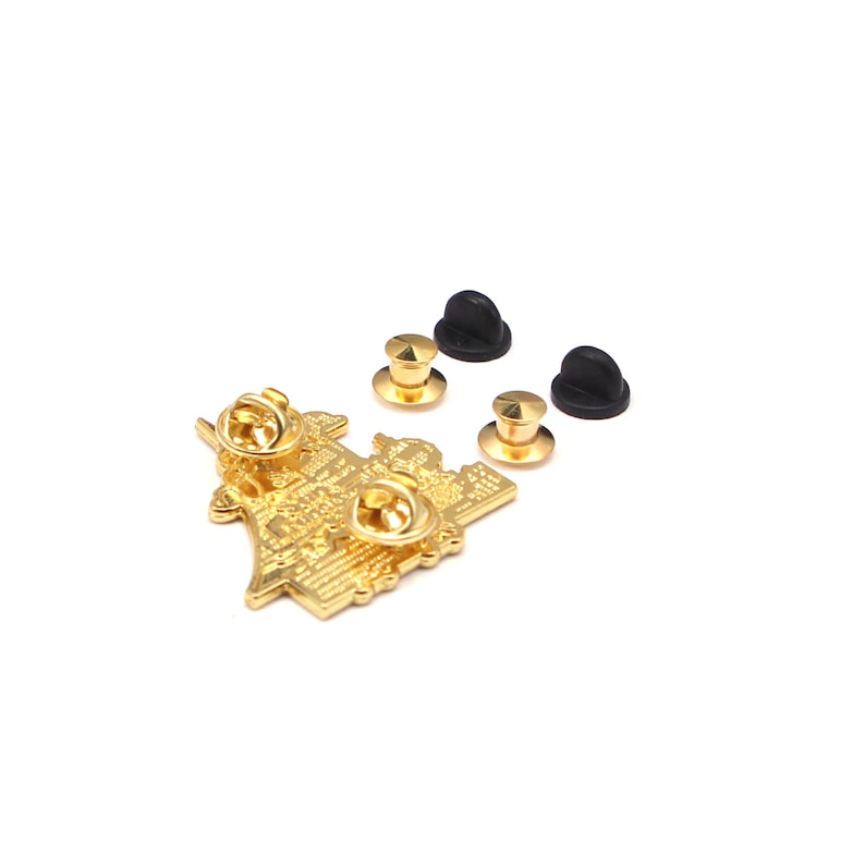 Op de afbeelding: Een goudkleurige metalen pin met gedetailleerd ontwerp en tekst. De pin heeft twee pin backs en wordt geleverd met drie goudkleurige pin clutches en drie zwarte rubberen clutches. De pin is geplaatst tegen een witte achtergrond.