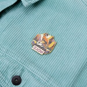 May include: A colorful enamel pin featuring a mountain ram and the text "ZION" on a teal corduroy jacket. The pin has a gold border and depicts a mountain scene with purple, orange, and yellow hues.