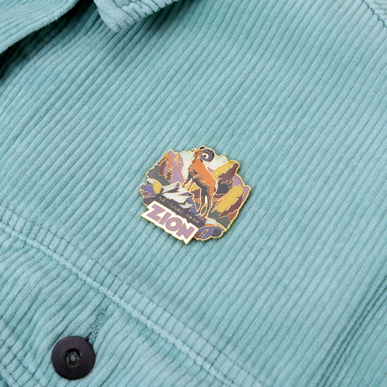May include: A colorful enamel pin featuring a mountain ram and the text "ZION" on a teal corduroy jacket. The pin has a gold border and depicts a mountain scene with purple, orange, and yellow hues.