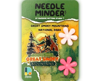 May include: A needle minder set featuring a pink flower design. The set includes a metal pin with a graphic of a bear and trees in front of a mountain range with the text "Great Smoky Mountains National Park".