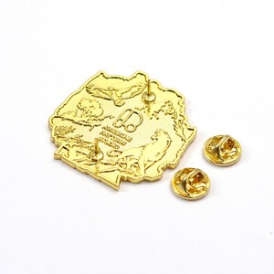 May include: A gold-colored pin with a detailed design, featuring an outline of a landmass with an eagle, tree, and other elements. Two additional gold pin backs are included. The pin has a shiny, reflective surface.