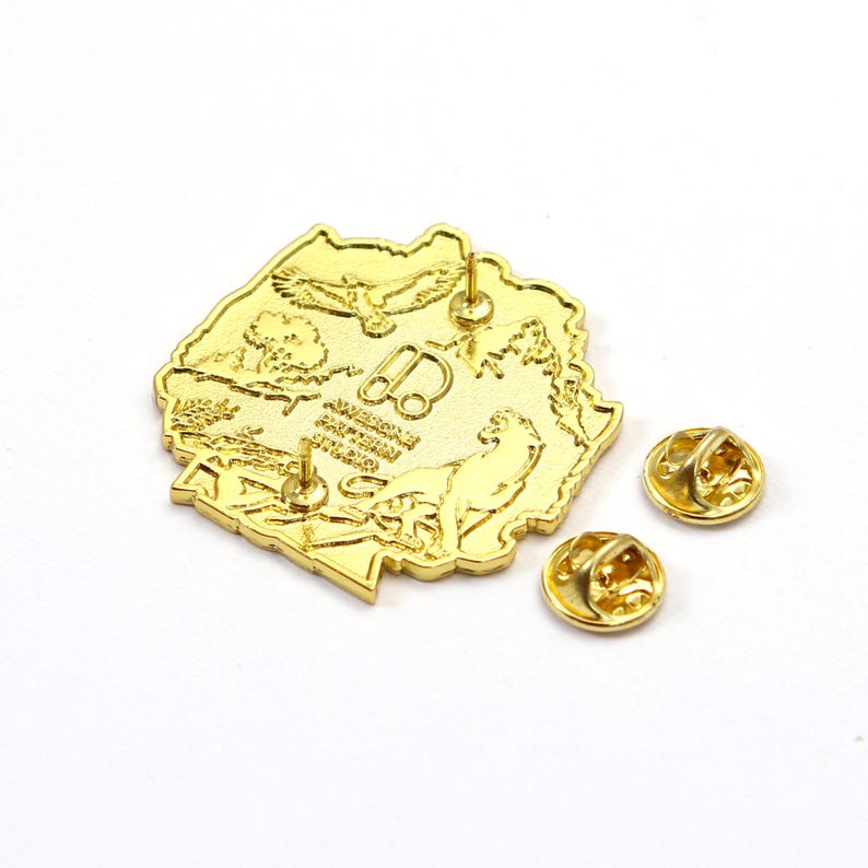 May include: A gold-colored pin with a detailed design, featuring an outline of a landmass with an eagle, tree, and other elements. Two additional gold pin backs are included. The pin has a shiny, reflective surface.