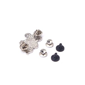 May include: A silver-colored metal pin with an ornate floral design, along with several pin backs. The pin has a textured surface and a classic aesthetic. The pin backs are silver and black.