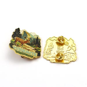 May include: Two Yosemite National Park enamel pins. One pin features a deer, trees, and mountains with the text "YOSEMITE NATIONAL PARK." The other pin is gold-colored with a mountain and tree design.