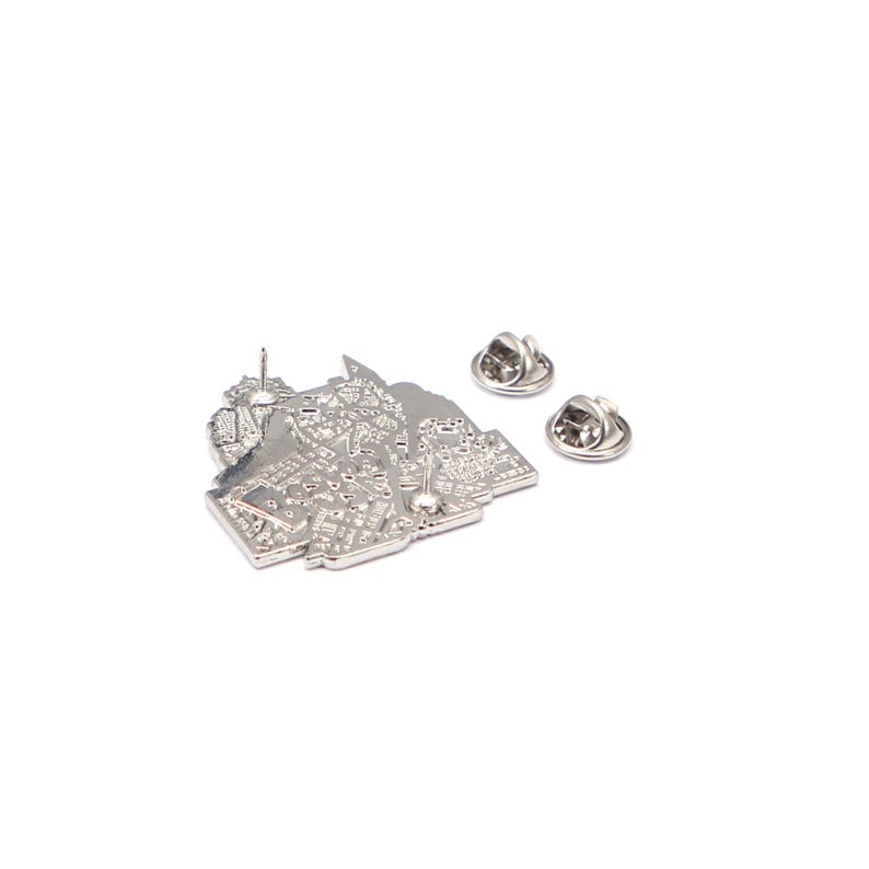 May include: A silver-coloured pin with a detailed city map design. The pin has a pin back and two additional pin backs are also visible. The pin is likely made of metal and is set against a white background.