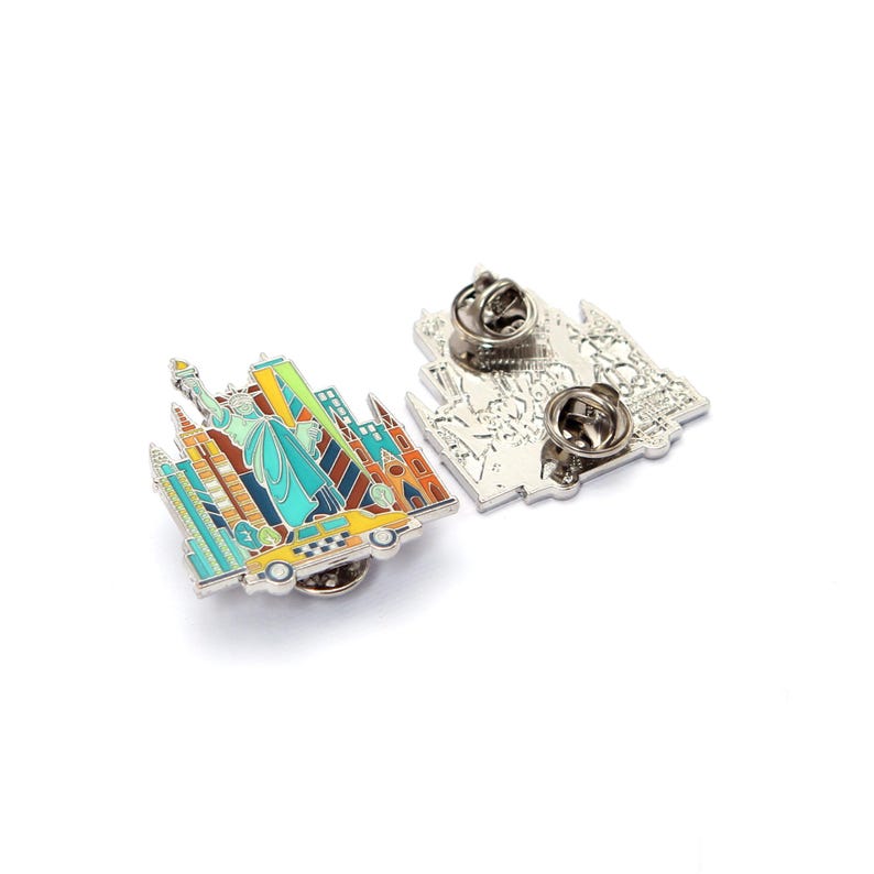May include: Two enamel pins featuring a New York City design. One pin shows the Statue of Liberty, skyscrapers, and a yellow taxi. The other pin shows the back of the design with the pin clasp.