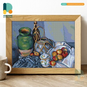Op de afbeelding: Kruissteekkunstwerk in een houten lijst. Het stilleven toont een groene vaas, een blauw-witte container, een kleine pot, een fles en een kom met rode appels. De achtergrond is een blauw-grijs patroon.