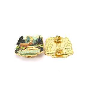 May include: A gold-colored Yosemite National Park enamel pin with a deer, trees, and mountains design. The pin features the text "YOSEMITE NATIONAL PARK" in green. The back of the pin is also gold-colored.