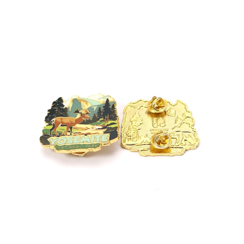 May include: A gold-colored Yosemite National Park enamel pin with a deer, trees, and mountains design. The pin features the text "YOSEMITE NATIONAL PARK" in green. The back of the pin is also gold-colored.