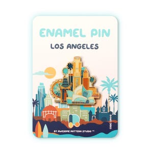 May include: Enamel pin featuring a Los Angeles cityscape design. The pin showcases iconic landmarks in shades of gold, blue, and orange, set against a backdrop with palm trees and a setting sun. The card reads "ENAMEL PIN LOS ANGELES".