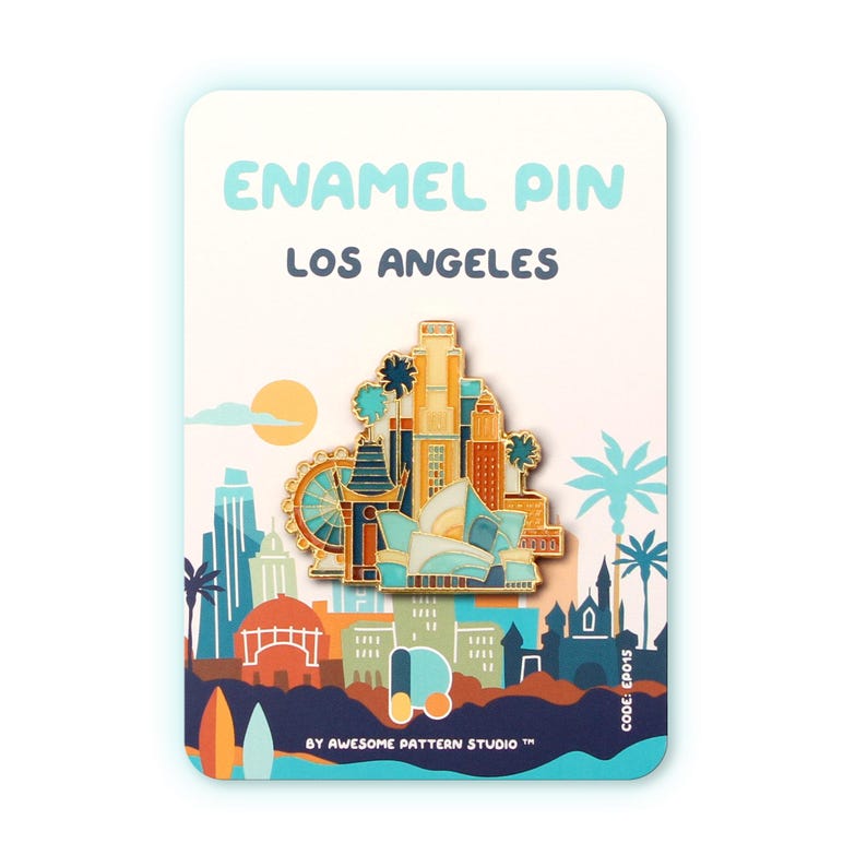 May include: Enamel pin featuring a Los Angeles cityscape design. The pin showcases iconic landmarks in shades of gold, blue, and orange, set against a backdrop with palm trees and a setting sun. The card reads "ENAMEL PIN LOS ANGELES".
