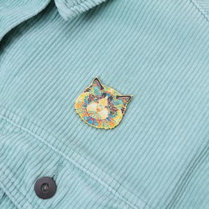 Puede incluir: Un pin decorativo con forma de gato y patrones intrincados en amarillo, azul y naranja. El pin est&aacute; colocado sobre una tela de pana azul claro, resaltando su dise&ntilde;o vibrante. Un accesorio de moda.