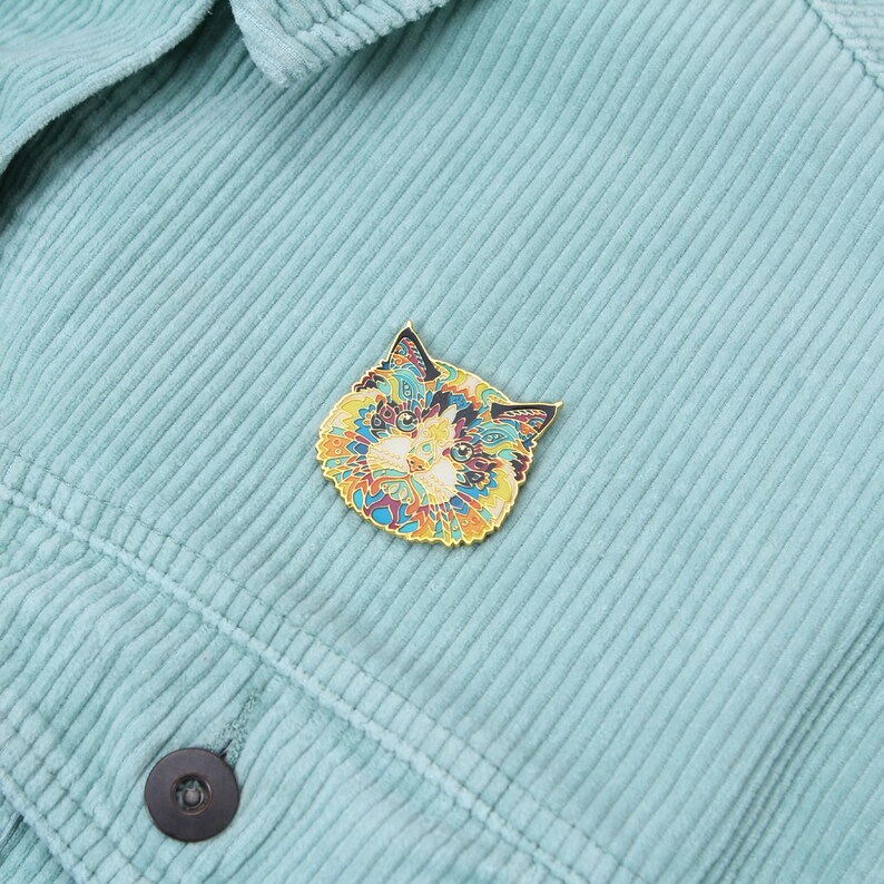 Puede incluir: Un pin decorativo con forma de gato y patrones intrincados en amarillo, azul y naranja. El pin est&aacute; colocado sobre una tela de pana azul claro, resaltando su dise&ntilde;o vibrante. Un accesorio de moda.