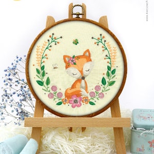 May include: Embroidered fox hoop art on a wooden easel. The fox is orange with white accents, wearing a floral crown, and surrounded by a floral wreath. The text "Awesome Pattern Studio with love" is in the upper left corner.