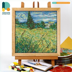 May include: A framed cross-stitch landscape featuring a field, trees, and sky, displayed on a wooden easel. The artwork is in a wooden frame. The scene is a vibrant mix of greens, blues, and yellows. The image includes the text "AWESOME PATTERN STUDIO".