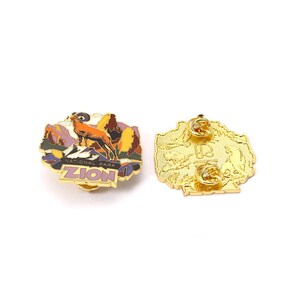 May include: A gold-colored enamel pin featuring a landscape scene with a mountain goat and the words "ZION NATIONAL PARK." The pin has a gold border and a butterfly clutch backing. The back of the pin is also gold.
