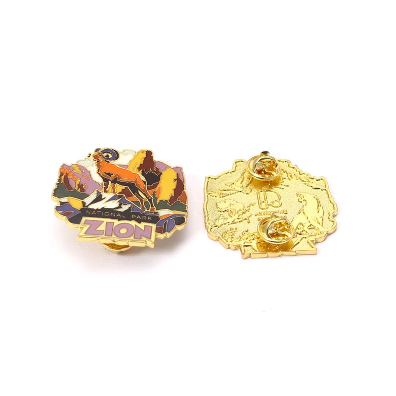 May include: A gold-colored enamel pin featuring a landscape scene with a mountain goat and the words "ZION NATIONAL PARK." The pin has a gold border and a butterfly clutch backing. The back of the pin is also gold.