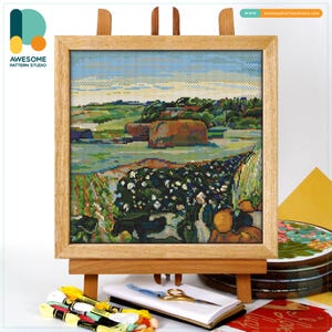 Può includere: Una scena di paesaggio a punto croce incorniciata in una cornice di legno, esposta su un cavalletto di legno. L'opera d'arte raffigura una campagna colorata con campi, alberi e uno specchio d'acqua. La scena è dettagliata con varie sfumature di verde, blu e marrone. L'indirizzo del sito web è visibile in alto a destra.