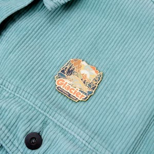May include: A Glacier National Park enamel pin with a detailed mountain and wildlife design, in shades of orange, blue, and green. The pin is attached to a teal corduroy jacket.