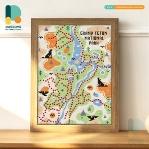 May include: A framed cross-stitch map of Grand Teton National Park, with a detailed trail layout in red, green, and blue. The map includes illustrations of wolves, trees, and other park features. The frame is made of light brown wood.