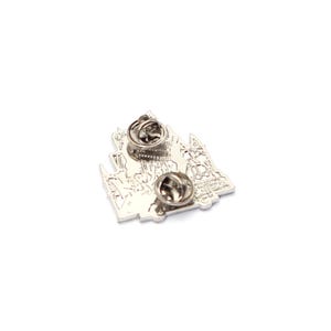 May include: A silver-colored metal pin with two pin backs. The pin features a detailed design of a building or castle, with intricate architectural elements. The pin is set against a white background.