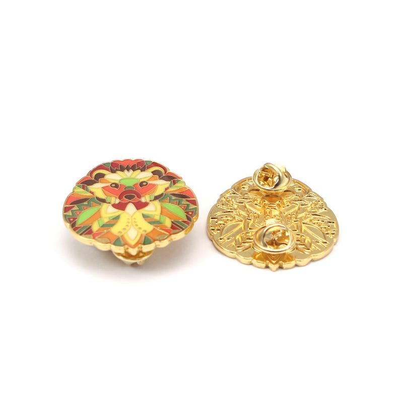 Puede incluir: Dos broches de esmalte dorados. Un broche presenta un dise&ntilde;o de erizo colorido con detalles en rojo, amarillo, verde y naranja. El otro broche muestra la parte posterior con un patr&oacute;n decorativo y un cierre de alfiler.