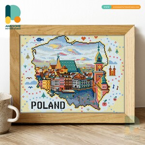 May include: A framed cross-stitch artwork showing a Polish cityscape within the country's outline. The image includes buildings, a church, and the word "POLAND". The frame is made of light-coloured wood.
