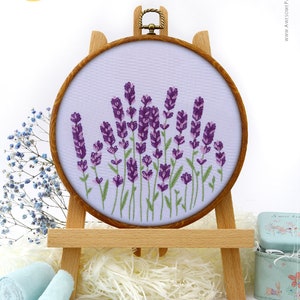 May include: A round embroidery hoop featuring a lavender design. The purple lavender flowers are stitched on a light purple fabric, set in a wooden hoop. The hoop is displayed on a small wooden easel. The image includes the text "Awesome Pattern Studio with love."
