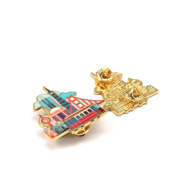 Op de afbeelding: Twee goudkleurige emaille pins met bezienswaardigheden van San Francisco. E&eacute;n pin toont de Golden Gate Bridge en een rode, blauwe en gouden kabeltram. De andere pin toont de achterkant met een naaldsluiting.