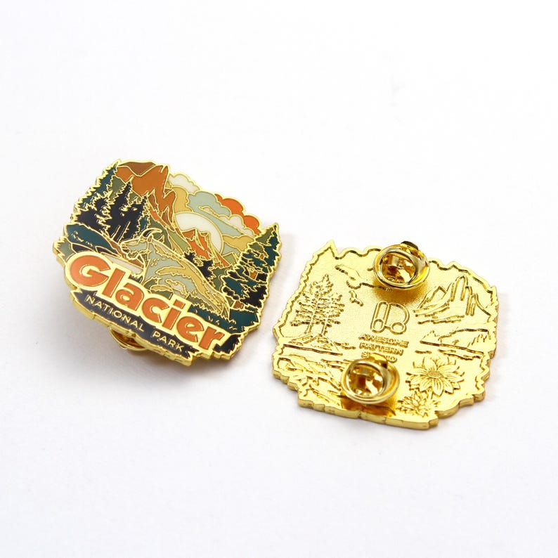May include: Two gold-colored enamel pins. One pin features a landscape design with the text "Glacier National Park." The other pin has a detailed landscape design with the text "Awesome Pattern Studio."