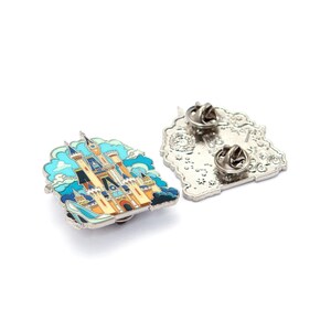 Pu&ograve; includere: Due spille smaltate con un design a castello colorato. Le spille raffigurano un castello delle fiabe con dettagli blu, gialli e arancioni, su uno sfondo di cielo blu e nuvole. Una spilla &egrave; mostrata con la chiusura e l'altra con la parte anteriore.
