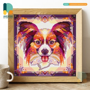 Op de afbeelding: Een kleurrijk kruissteekpatroon van een Papillon-hond met een paarse achtergrond. De vacht van de hond is een mix van oranje, rood en geel, met zwarte accenten. De ogen van de hond zijn bruin en zijn tong steekt uit. Het patroon is ingelijst in een houten lijst.