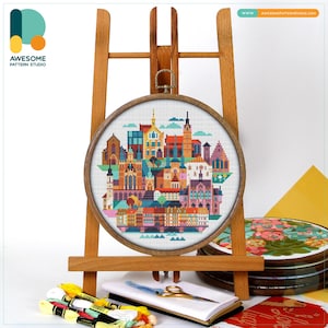 Peut inclure: Un tambour à broder circulaire en bois contenant un motif au point de croix représentant une ligne d'horizon urbaine européenne colorée avec divers bâtiments, tours et maisons dans des tons orange, bleu, rose et jaune, sur un chevalet.