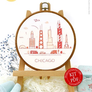 Op de afbeelding: Een kruissteek-borduurring met een witte achtergrond en een rood-wit ontwerp van de skyline van Chicago. De ring staat op een houten standaard en de tekst "CHICAGO" is onder het ontwerp gedrukt. De afbeelding bevat ook een rode cirkel met de tekst "CROSS-STITCH PATTERN KIT & PDF CUSTOMIZABLE".