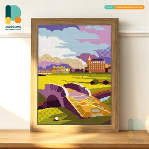 May include: A framed cross-stitch landscape depicts a golf course with a bridge, buildings, and mountains under a blue and purple sky. The artwork is in a wooden frame and sits on a wooden surface.