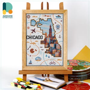 May include: A framed cross-stitch art piece depicting the Chicago skyline and city map. The artwork features the word "CHICAGO" in bold letters, with a color palette of blue, orange, and brown. The frame is made of light-colored wood, and the piece is displayed on a wooden easel.
