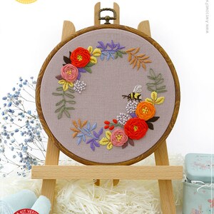 May include: An embroidery hoop featuring a floral and bee design. The design includes red, orange, pink, yellow, and purple flowers, green leaves, and a bumblebee. The embroidery is set on a gray fabric and displayed on a wooden easel.