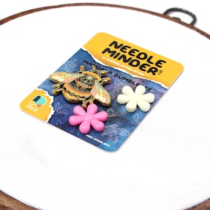 May include: A needle minder set featuring a gold-colored bee pin with colorful details, two flower-shaped magnets, and a card with the text "NEEDLE MINDER". The bee pin is approximately 1 inch long. The set is designed for embroidery and cross-stitch projects.
